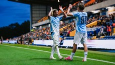 Two football players in blue jerseys celebrating on the sideline of a stadium during a match, raising their hands and smiling, with fans in the background.