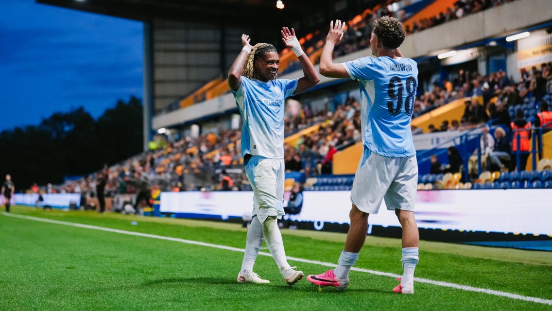 Two football players in blue jerseys celebrating on the sideline of a stadium during a match, raising their hands and smiling, with fans in the background.