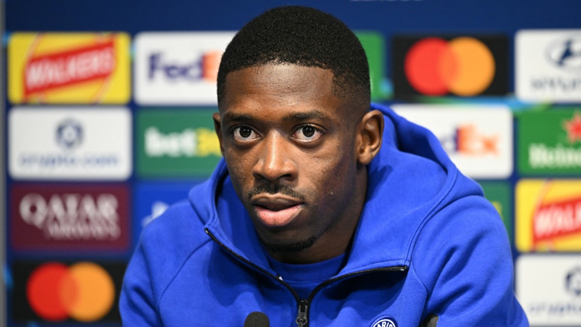 A soccer player in a blue jacket at a press conference with a backdrop of sponsor logos.
