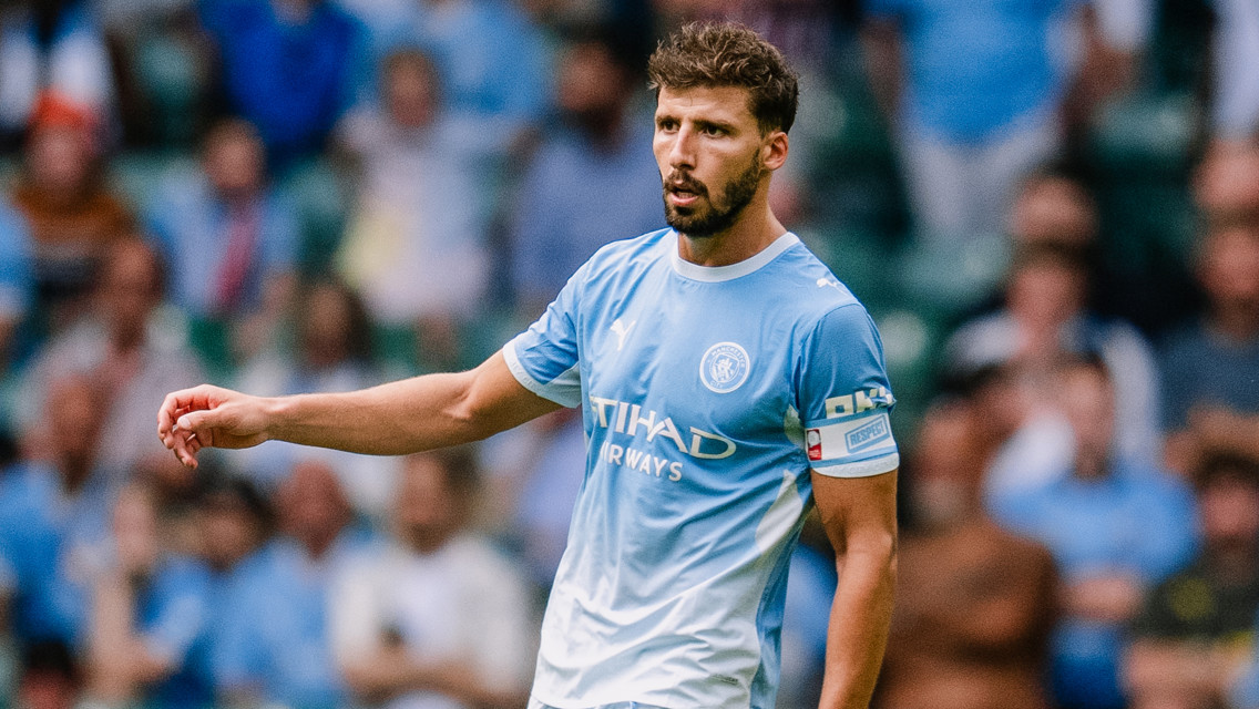 A Manchester City player wearing a light blue jersey with the Etihad Airways logo, standing on the field and gesturing with his hand.