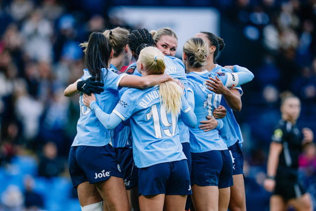 A group of women soccer players wearing blue jerseys with names and numbers are huddled together in celebration on a soccer field.