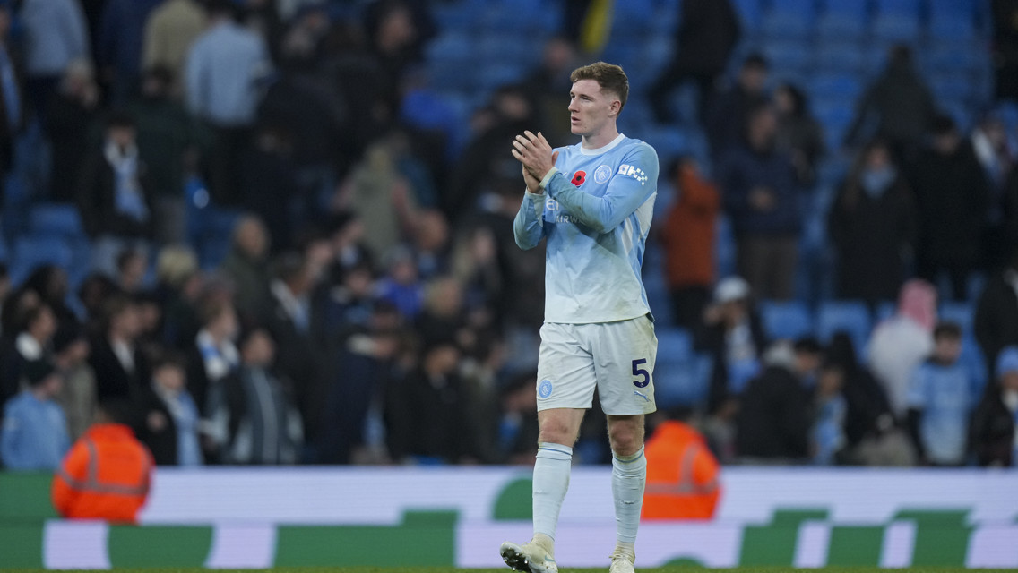 A Manchester City player clapping on the field at the Etihad Stadium with fans in the background.