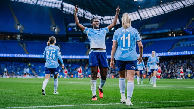 Manchester City women's soccer team celebrating a goal at the Etihad Stadium. Players wearing blue kits with numbers 25 and 11 visible.