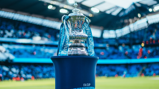 The image shows the FA Cup trophy on a display stand with ribbons labeled 'Emirates Fly Better.' It is situated in a stadium, with blurred spectators and seating in the background.