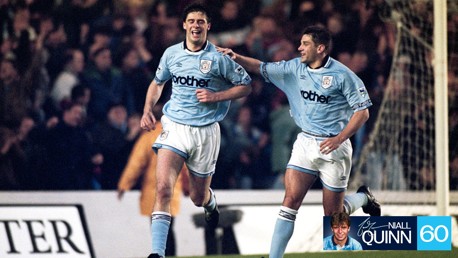 Two soccer players in Manchester City jerseys celebrate on the field. The player on the left, with a broad smile, runs with arms raised. The other places a hand on his teammate's back.