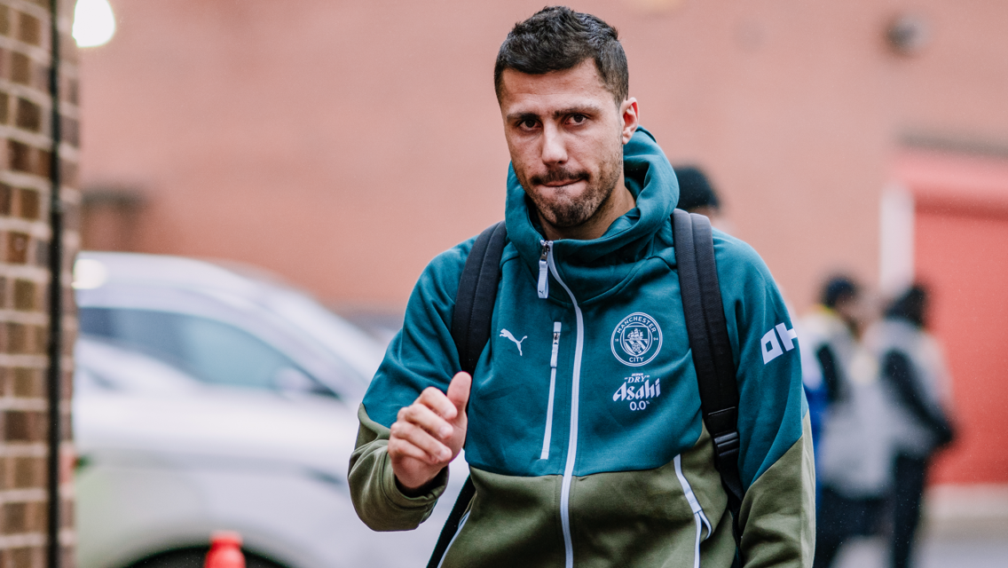 A person wearing a Manchester City tracksuit and carrying a backpack walks outdoors. The tracksuit has the Manchester City and Puma logos.