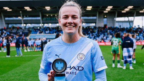 A Manchester City player holds a trophy on a soccer field with celebrating teammates in the background.