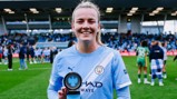 A Manchester City player holds a trophy on a soccer field with celebrating teammates in the background.