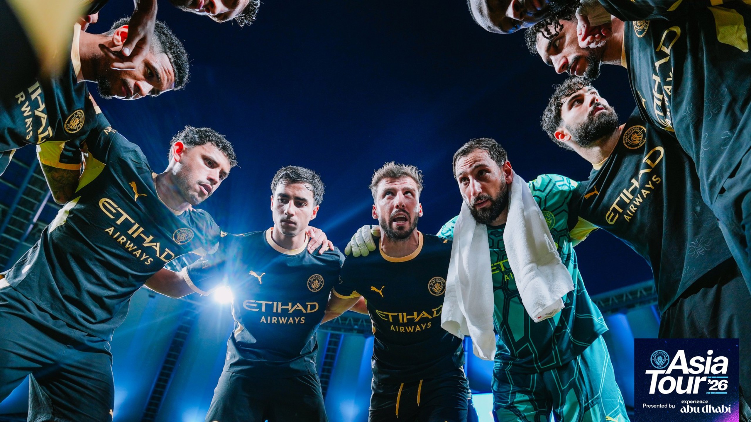 Players in Manchester City jerseys huddling with 'Asia Tour '26' branding. The jerseys display the Etihad Airways logo.