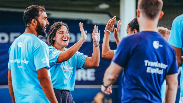 A group of people wearing light blue shirts, participating in a team-building exercise. They are giving high-fives to each other and the shirts have the text Young Leader and Translator Español on them.