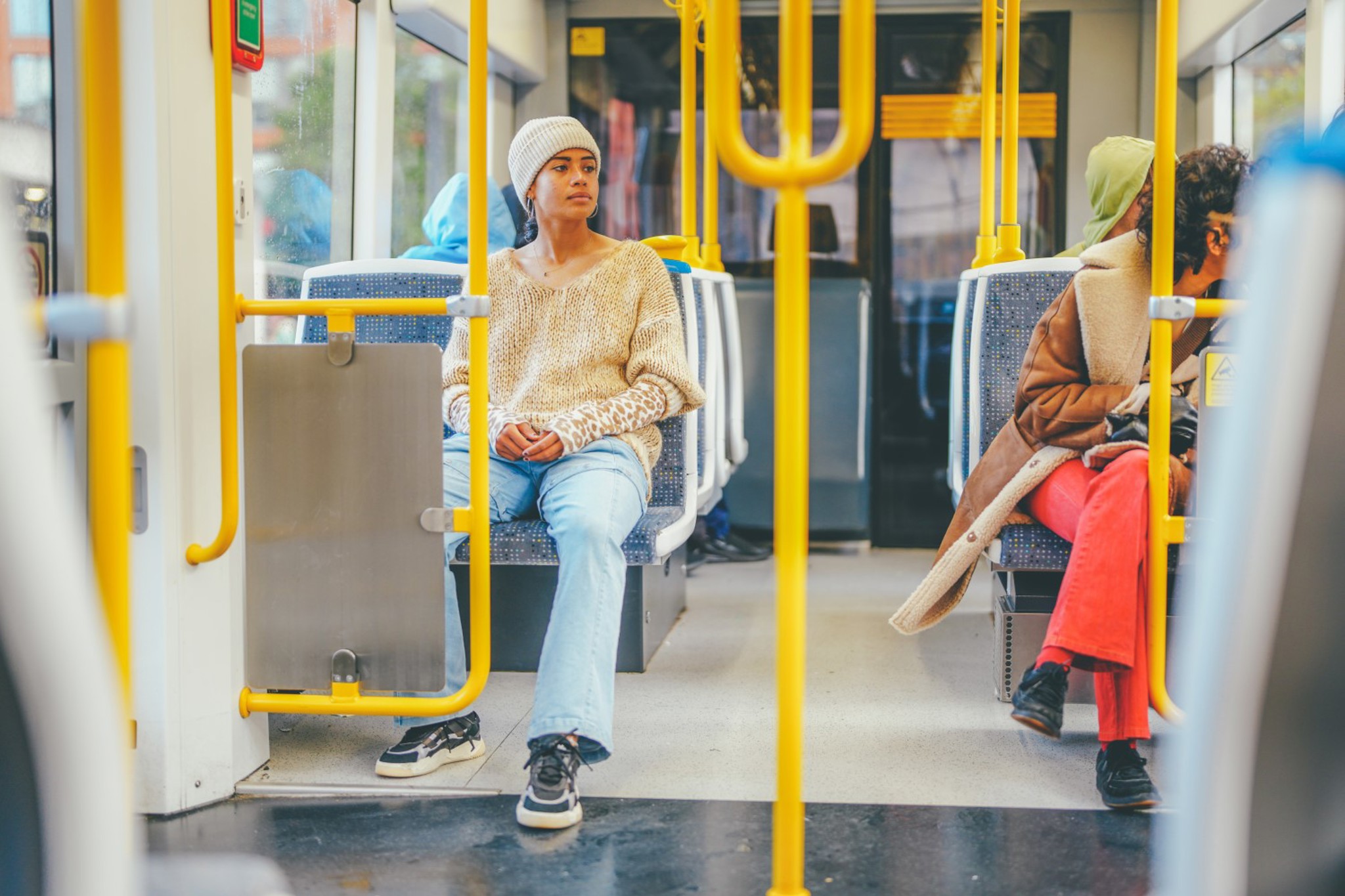 Three passengers sitting on a train with yellow poles and grey seating.