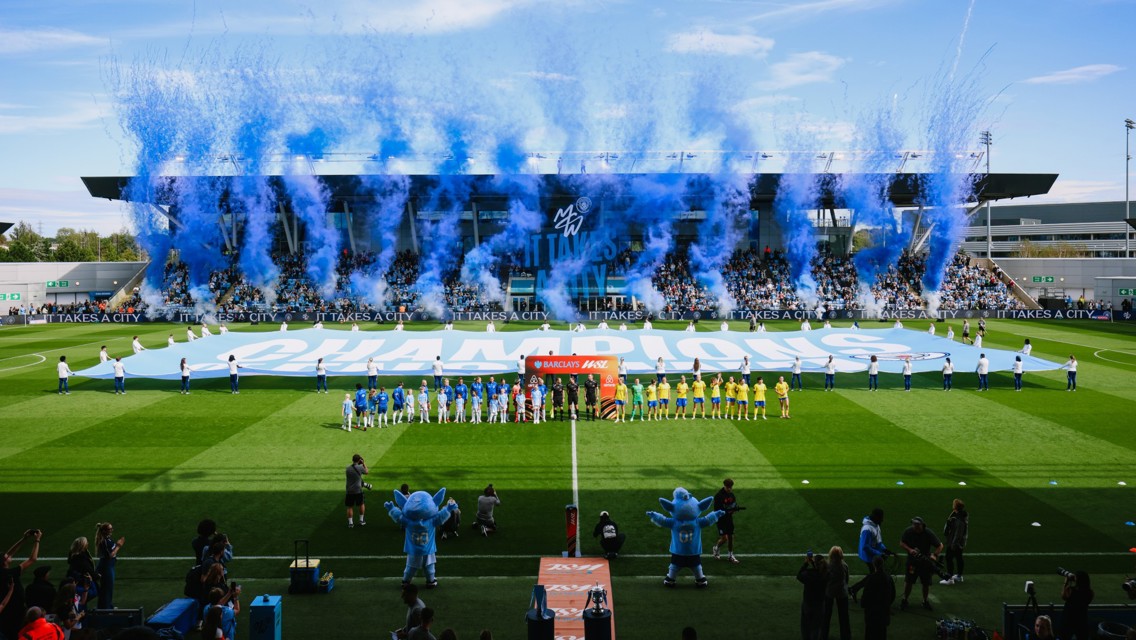 Manchester City WSL teams line up on the field with blue confetti and smoke celebrating a championship. Large banner reading 'Champions' displayed.