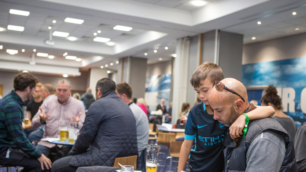 A crowded room with people socializing; a boy wearing a Manchester City jersey leans on a man checking his phone.