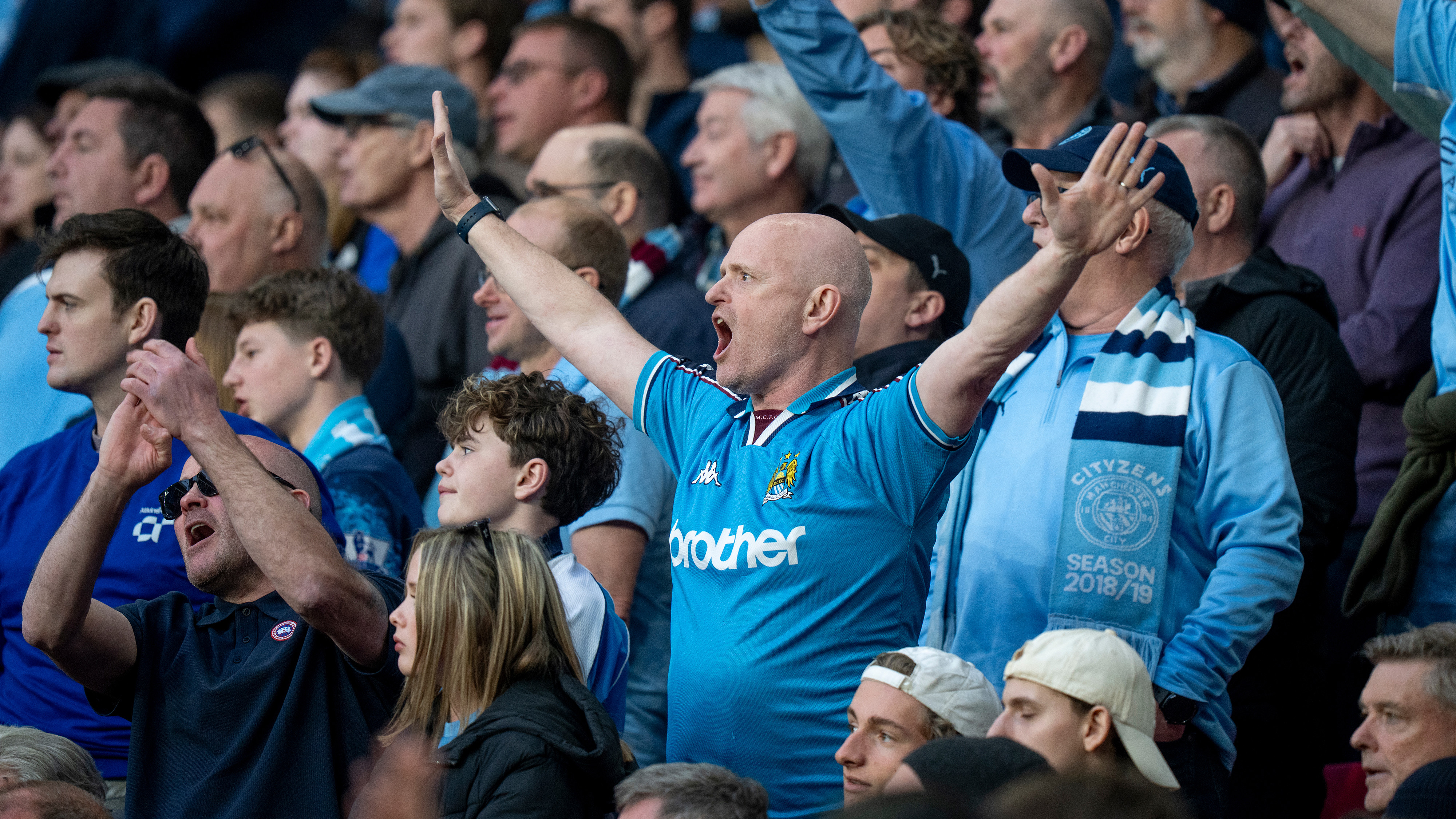 Fans in blue jerseys cheering during a football match, some wearing scarves, in a crowded stadium.