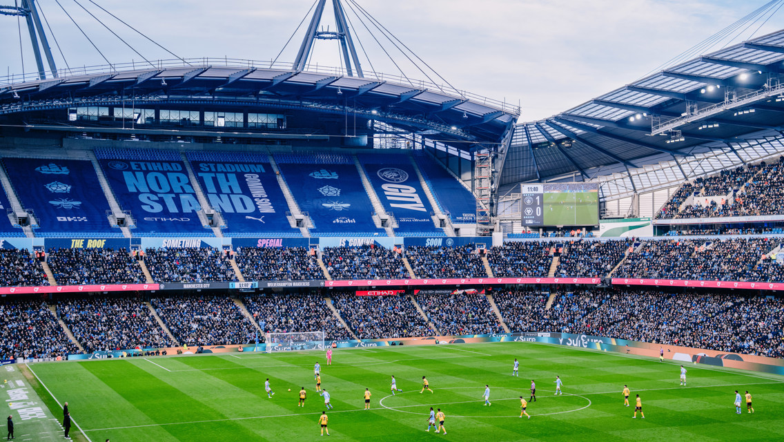 A football match taking place at Etihad Stadium, spectators in the stands watching the game unfold on the field.