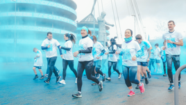 Participants in a fun run event pass through blue color smoke near a stadium with a visible statue and text 'Official Supporters Clubs' in the background.