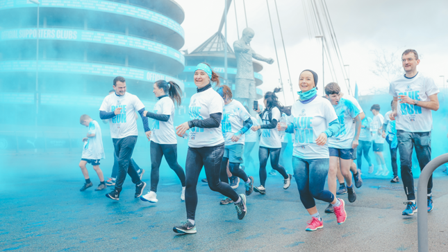 Participants in a fun run event pass through blue color smoke near a stadium with a visible statue and text 'Official Supporters Clubs' in the background.