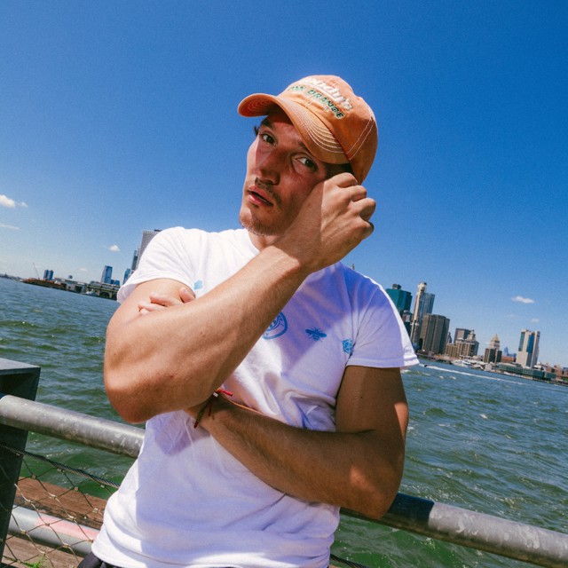 A person wearing a white shirt and orange cap stands by a waterfront railing with the city skyline visible in the background.
