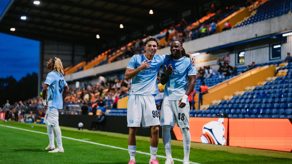 Two Manchester City players celebrating on the field with a player in the background at a stadium.