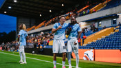 Two Manchester City players celebrating on the field with a player in the background at a stadium.