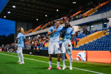 Two Manchester City players celebrating on the field with a player in the background at a stadium.