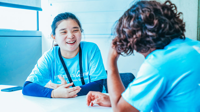 Two individuals are seated across from each other at a table, engaged in conversation. Both are wearing blue shirts, and a mobile phone is placed on the table beside them. The faces are blurred.