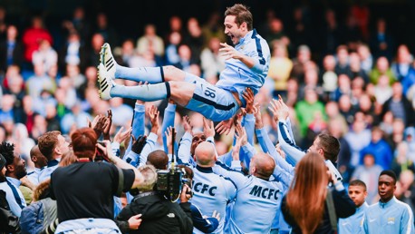 A football team in light blue kits celebrates a player by lifting him in the air. The player wears jersey number 18. Team members and photographers surround him in a stadium environment.