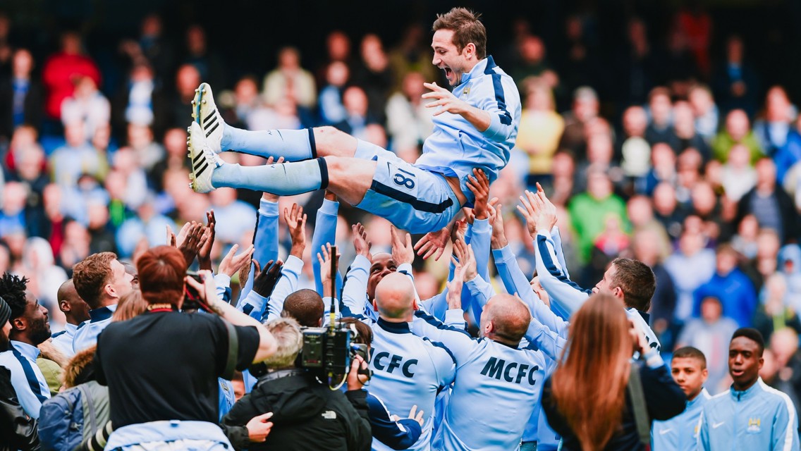 A football team in light blue kits celebrates a player by lifting him in the air. The player wears jersey number 18. Team members and photographers surround him in a stadium environment.