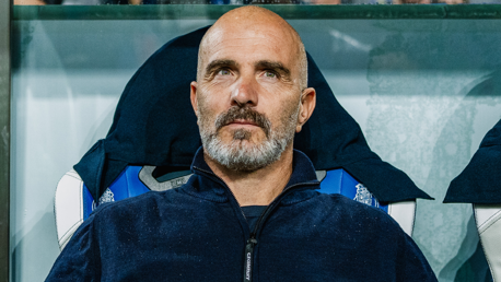 A bald man with a beard wearing a dark jacket is seated, possibly during a sports event.