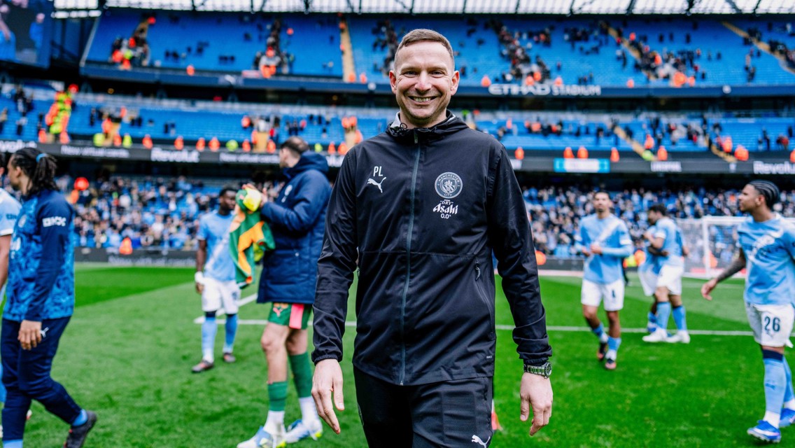 A person in a Manchester City tracksuit is walking on the pitch at the Etihad Stadium with players in the background.