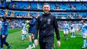 A person in a Manchester City tracksuit is walking on the pitch at the Etihad Stadium with players in the background.