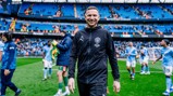 A person in a Manchester City tracksuit is walking on the pitch at the Etihad Stadium with players in the background.