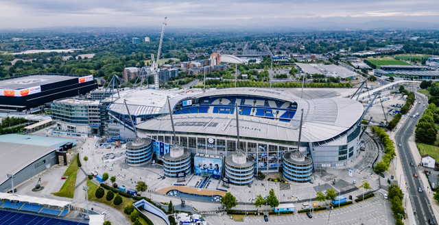 Aerial view of the Etihad Stadium in Manchester, with surrounding buildings and roads visible.