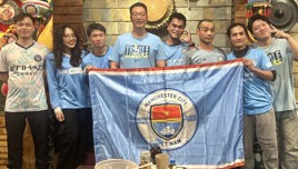 Group of people wearing Manchester City jerseys holding a Manchester City Vietnam flag indoors.