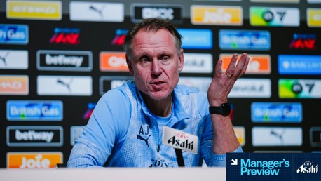 A manager preview press conference with a blurred face. The person is wearing a light blue jacket with 'AJ' on it. The backdrop features various logos, including Puma, Betway, and British Gas.