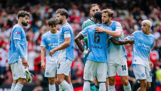 A group of Manchester City football players in light blue jerseys huddling on the field during a match.