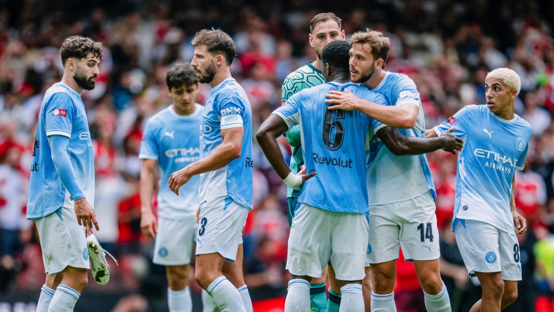 A group of Manchester City football players in light blue jerseys huddling on the field during a match.