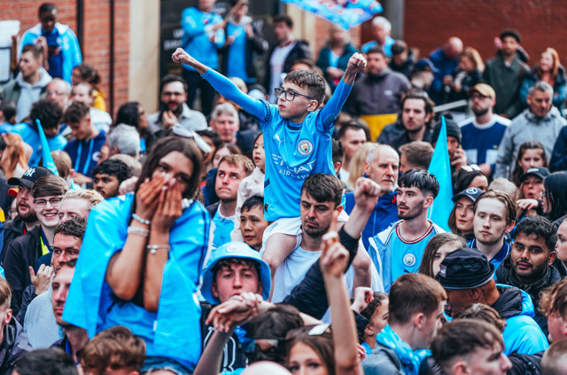 A crowd of Manchester City fans celebrating with flags and scarves. A child on an adult's shoulders in a blue jersey cheers enthusiastically.