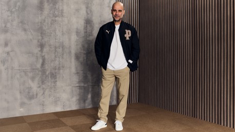 Person wearing a black Puma jacket, white shirt, beige pants, and white shoes, standing against a gray and wooden background.