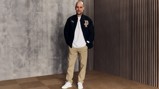 Person wearing a black Puma jacket, white shirt, beige pants, and white shoes, standing against a gray and wooden background.