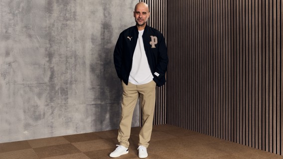 Person wearing a black Puma jacket, white shirt, beige pants, and white shoes, standing against a gray and wooden background.