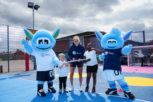 At a community event, two people dressed as Manchester City mascots, Moonchester and Moonbeam, accompany children and an adult holding up a ceremonial ribbon on a sports pitch.