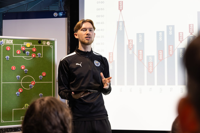 A soccer coach presenting tactics on a field board and a chart, wearing a Manchester City tracksuit.