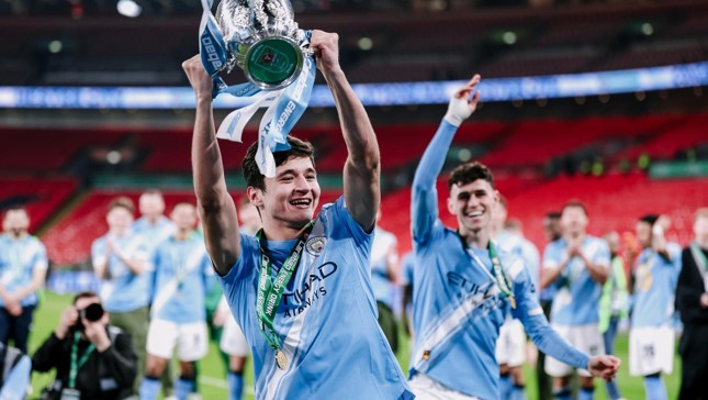 Football players wearing Manchester City jerseys celebrating with a trophy on the field.