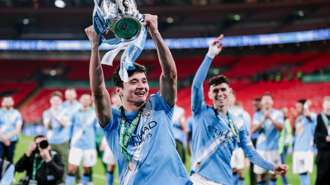 Football players wearing Manchester City jerseys celebrating with a trophy on the field.