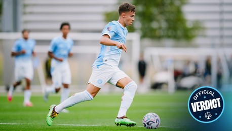 A soccer player in a light blue kit, running with the ball on a field during an intense match. Teammates blur in the background, focused on the game.