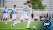 A soccer player in a light blue kit, running with the ball on a field during an intense match. Teammates blur in the background, focused on the game.