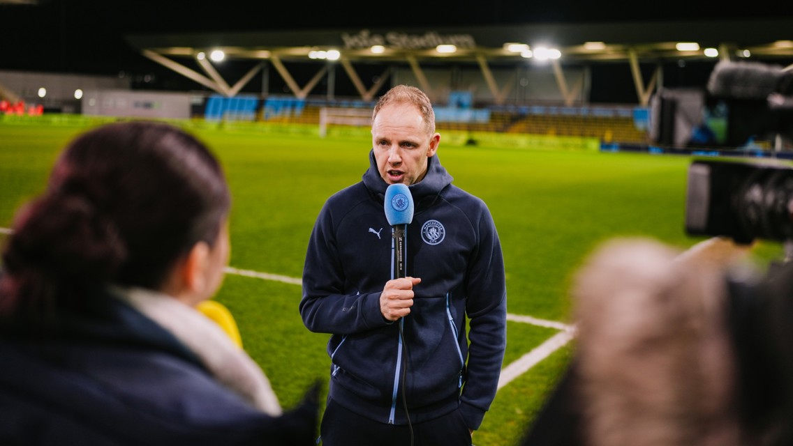 Person in a Manchester City jacket giving an interview on a football pitch with recording equipment around.