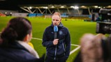 Person in a Manchester City jacket giving an interview on a football pitch with recording equipment around.
