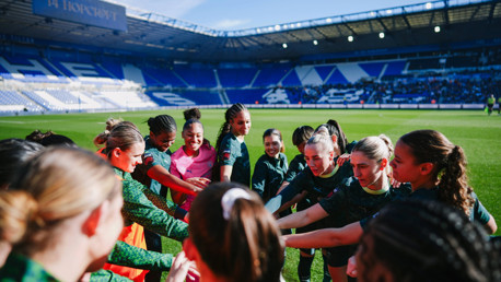 A group of football players in green jerseys huddled together on a stadium field with empty stands in the background.